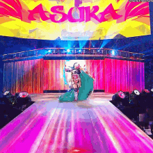 a woman in a green cape is walking down a stage with a sign that says asura