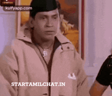 a man wearing a hat and a jacket is standing in front of a wall with a sign that says starttamilchat.in