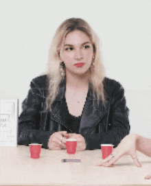 a woman in a leather jacket sits at a table with three red cups on it