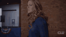 a woman in a superman costume stands in front of a crowd in a room with a cw logo on the wall
