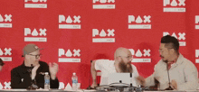 three men are sitting at a table in front of a red wall that says pax east