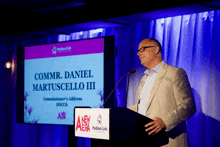 a man stands at a podium with a screen behind him that says commr daniel martuscello iii
