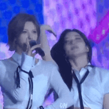 two women are standing next to each other on a stage . one of the women is singing into a microphone .