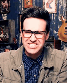 a man wearing glasses and a jacket smiles in front of a guitar