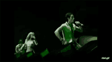 a man is singing into a microphone on a stage while a woman stands behind him .