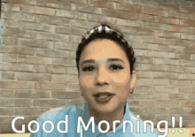 a woman in a blue shirt says good morning in front of a brick wall .