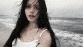 a woman with long black hair is standing on the beach looking at the camera .