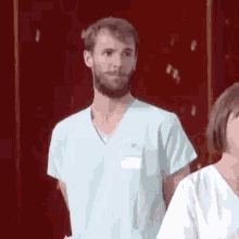 a man with a beard is wearing a scrub top and standing next to a woman in a white shirt .