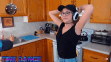 a woman in a kitchen wearing headphones and a black tank top with the word trick dance on the bottom right