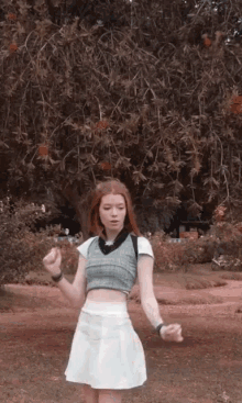 a girl wearing a crop top and a white skirt stands in front of a tree