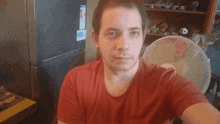 a man in a red shirt stands in front of a fan and a refrigerator