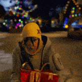 a person wearing a yellow hat with an x on it is holding a red gift in front of a christmas tree