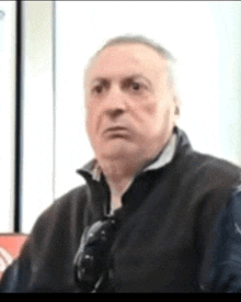 an older man wearing sunglasses and a black vest is looking at the camera .