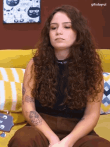 a woman with curly hair and a tattoo on her arm is sitting on a yellow couch .