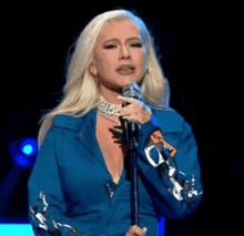 a woman in a blue jacket singing into a microphone with a sticker on her sleeve that says ' x '