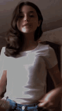 a woman wearing a white t-shirt and blue jeans is standing in front of a wall