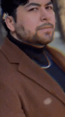 a man with a beard wearing a brown coat and black shirt