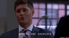 a man in a suit and tie is saying `` strike one sherlock '' while talking to another man .