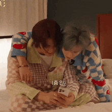 two people sitting on a bed looking at a cell phone with korean writing on the bottom