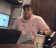a man in a pink shirt is sitting at a desk with a cup of coffee and a laptop .