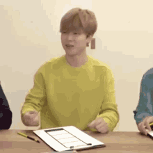 a young man in a yellow sweater is sitting at a table with a clipboard and pen .