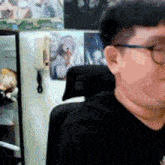 a man wearing glasses and a black shirt is sitting in a chair in front of a wall with pictures on it