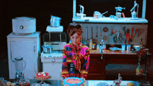 a woman in a colorful shirt is standing in a kitchen with a cake on the table