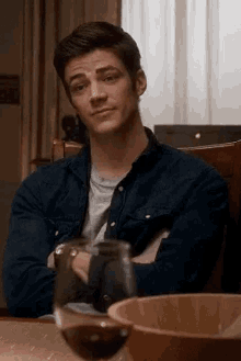 a man in a denim shirt is sitting at a table with his arms crossed and a glass of wine .