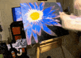 a painting of a blue flower with a yellow center is on an easel