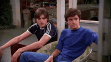two men are sitting next to each other on a porch and one is wearing a blue shirt