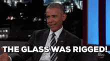 a man in a suit and tie is sitting on a couch with the words " the glass was rigged " behind him .