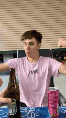 a young man in a pink shirt is sitting at a table with a bottle of beer and a can of blut