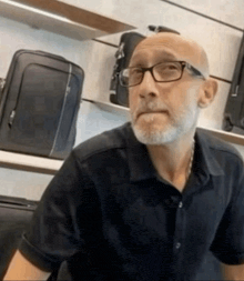 a bald man with glasses and a beard is sitting in front of a shelf of luggage