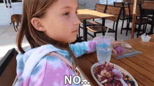 a little girl sitting at a table with a plate of food and a cup of water with the word no written on it