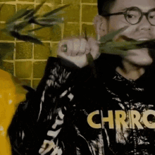 a man wearing glasses and a chrro shirt is holding a plant in his mouth .