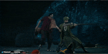 a man in a turban is holding a rifle in front of another man in a red shirt