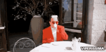 a man in a red suit is sitting at a table drinking a cup of coffee with the words viperissima above him