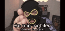 a man wearing a mask and headphones points at the camera with the words ge mig mobiltelefon ( give me the phone ) below him