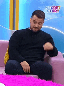 a man in a black sweater is sitting on a couch with a secret story logo in the background .