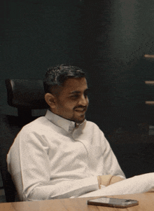 a man in a white shirt sits at a desk with a cell phone on the table