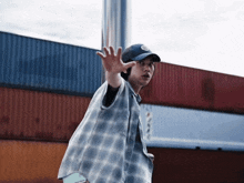 a man wearing a plaid shirt and a baseball cap is standing in front of a row of shipping containers