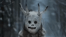a person wearing a white mask with horns on their head is standing in the woods .