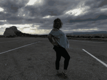 a woman standing on the side of a road with a cloudy sky in the background