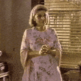 a woman in a floral dress is standing in front of a window with blinds