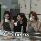 three women sit at a table drinking coffee and the word cheers is displayed