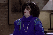 a woman in a purple sweater and glasses is sitting on a couch with her hands on her chest .