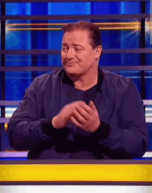a man in a blue jacket is sitting at a table with his hands folded and smiling .