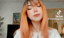 a woman with orange hair and bangs is wearing a white shirt and looking at the camera .