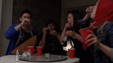 a group of people are sitting around a table with shot glasses and red cups