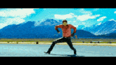 a man in a red shirt is dancing on the side of a road with mountains in the background